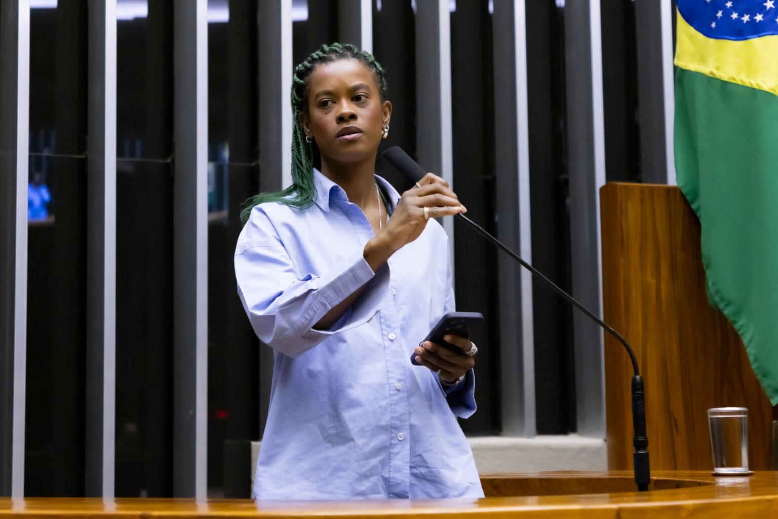 Deputada está discursando em plenário. Ela veste uma camisa de manga longa azul marinho. Atrás dela tem parte da bandeira do Brasil